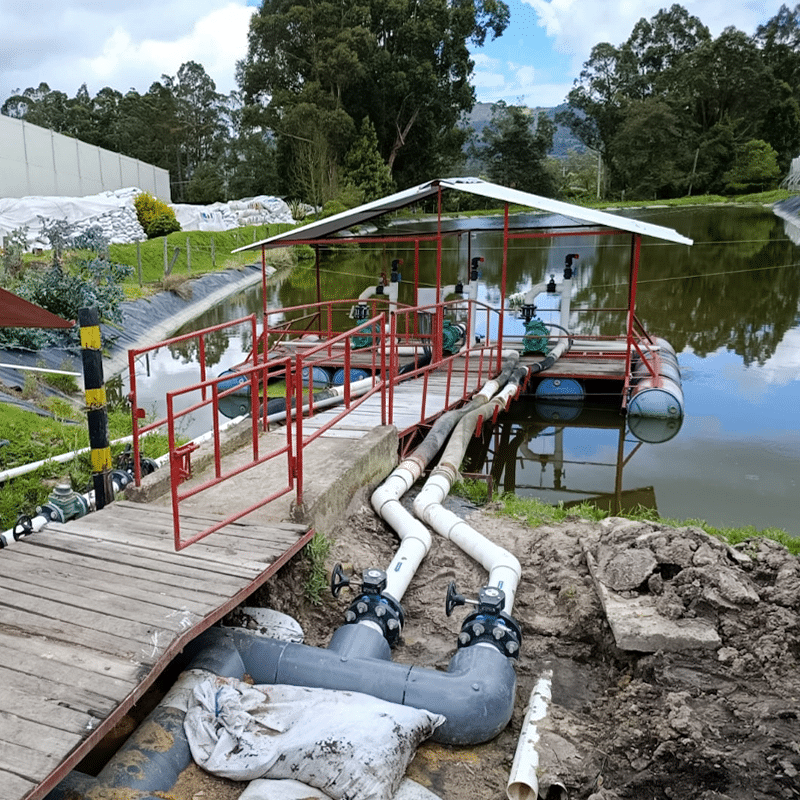 Water treatment setup with a red railing platform over a pond, large white and gray pipes, and surrounding greenery.