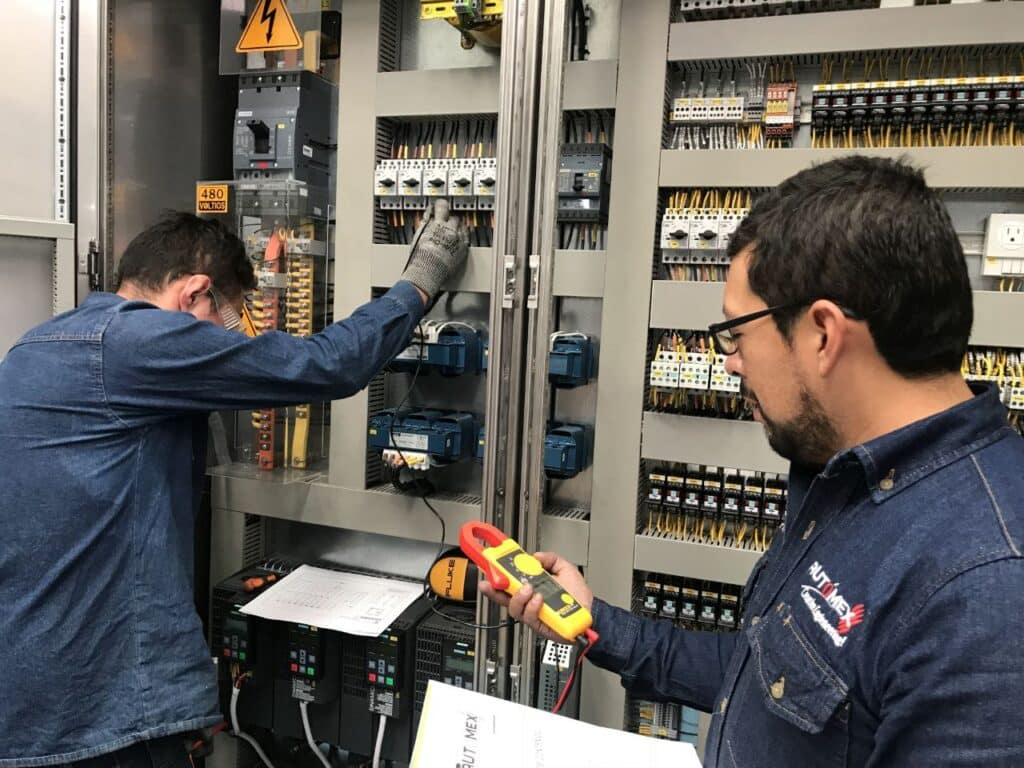Two technicians in denim shirts inspect and test an open electrical control panel, one using a yellow Fluke multimeter while the other works inside the panel with gloves on.