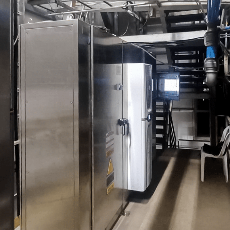 Industrial equipment room: a large stainless steel control cabinet with a white cooling unit beside it, overhead pipes, and a small monitor on a stand in the background.