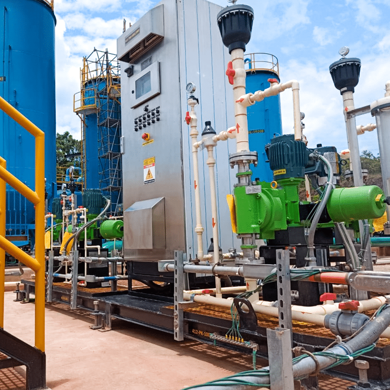Outdoor industrial facility with a control cabinet, blue storage tanks, and interconnected white pipes and green pumps on a metal platform.