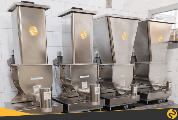 Four stainless-steel bulk ingredient dispensers with conical hoppers lined up on a counter in a clean food-processing facility, each bearing a yellow logo.