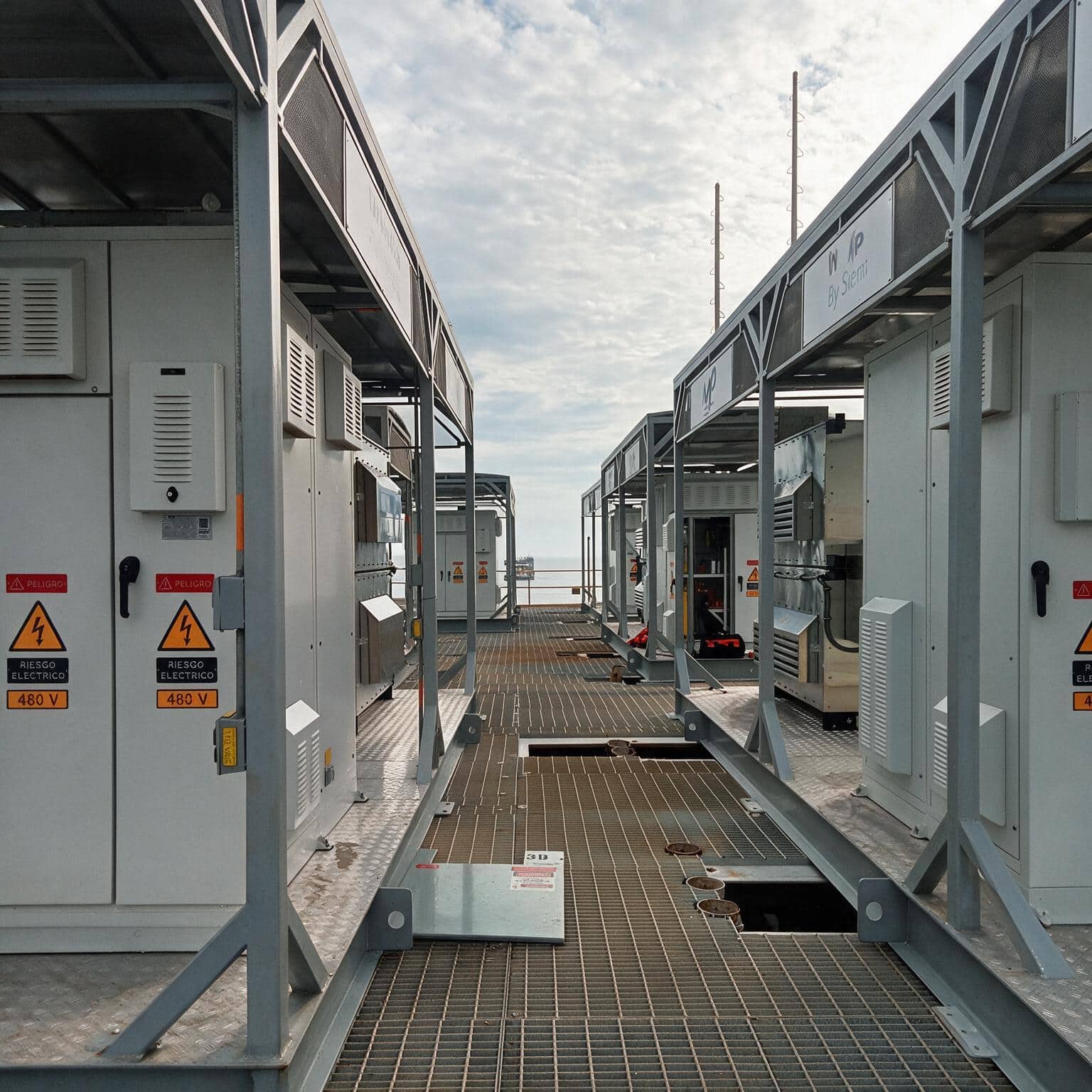 Industrial outdoor catwalk between rows of grey electrical cabinets with warning labels and a metal grating floor, led toward the horizon over water.