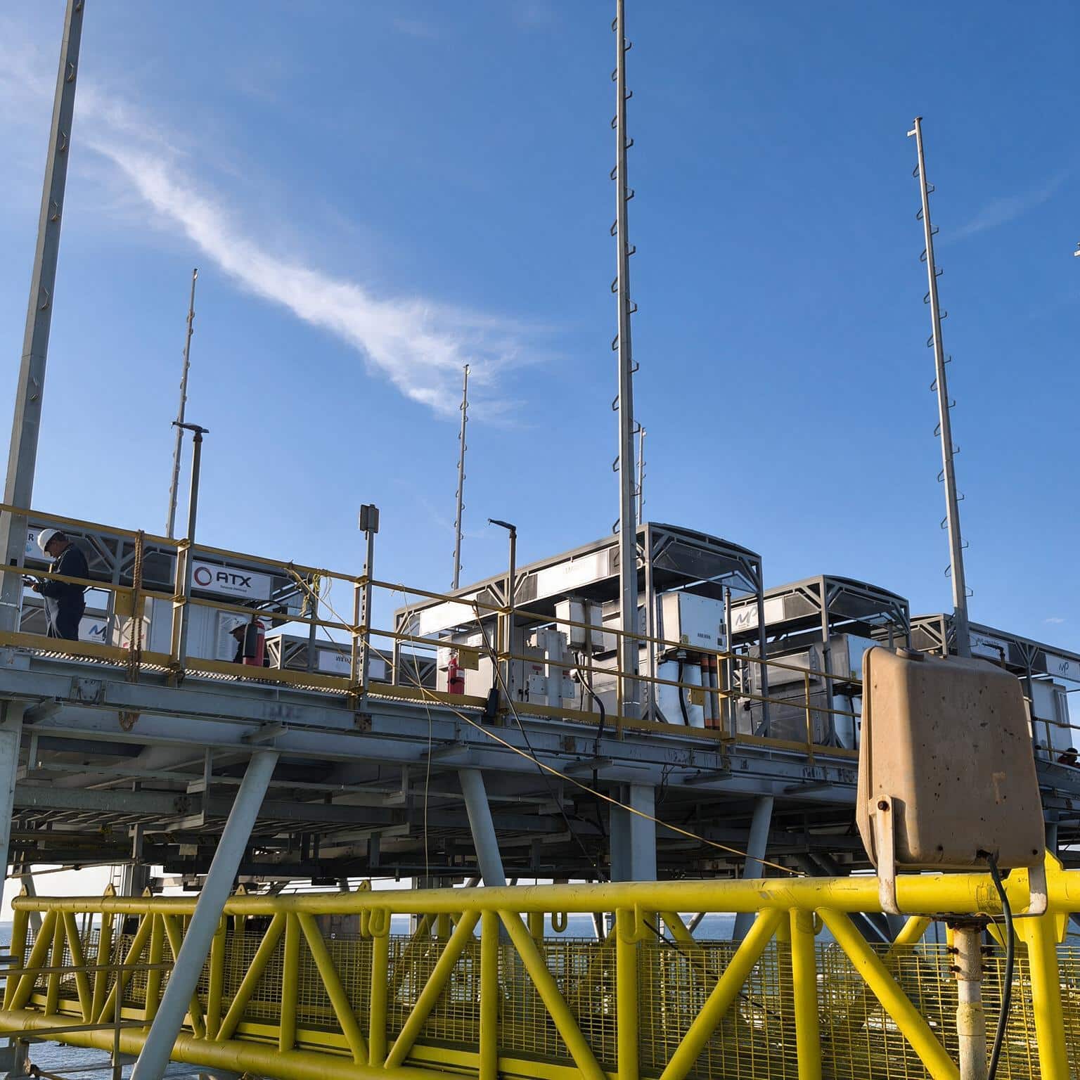 Industrial platform with yellow safety rails and metal walkways, tall vertical masts against a blue sky, worker in a hard hat on the left.